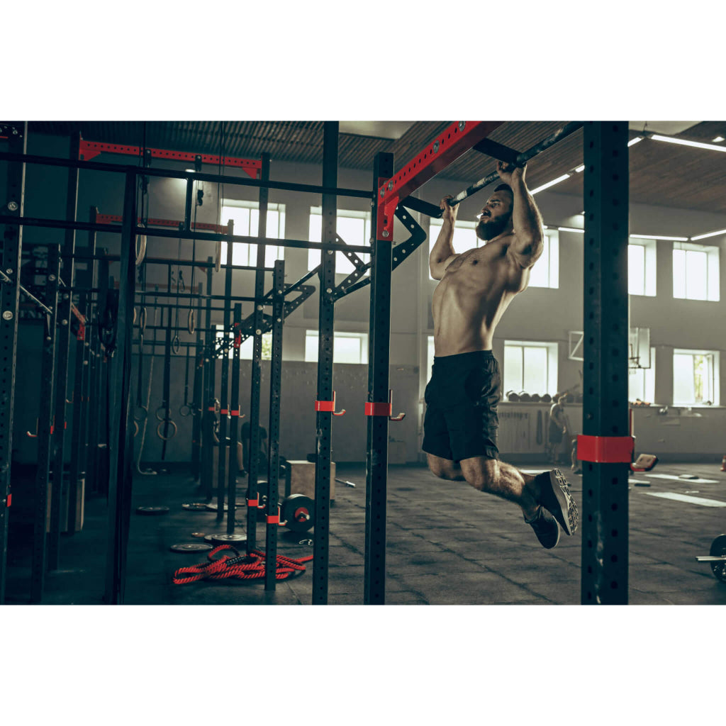 Main Website photo of a muscular man working out on a red and black power rack doing pull ups